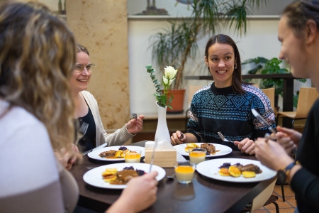 Vier Personen sitzen an einem kleinen Tisch mit Tellern voller Essen und kleinen Gläsern mit einer Nachspeise in einem hellen Raum mit Pflanzen im Hintergrund.