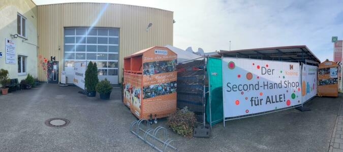 Außenbereich mit einem großen Banner 'Der Second-Hand-Shop für ALLE!' neben großen orangefarbenen Kleidercontainern und einem Gebäude mit großen Fenstern.