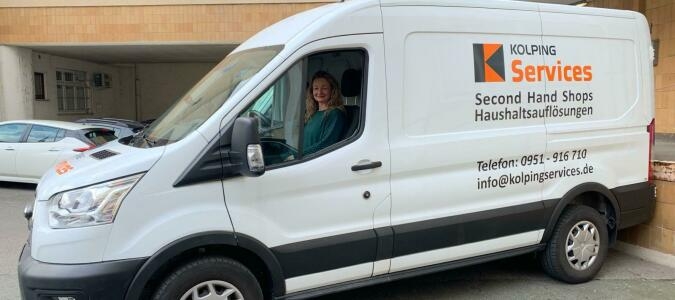 Weißer Transporter mit der Aufschrift 'KOLPING Services Second Hand Shops Haushaltsauflösungen' und Kontaktdaten, geparkt vor einem Gebäude.