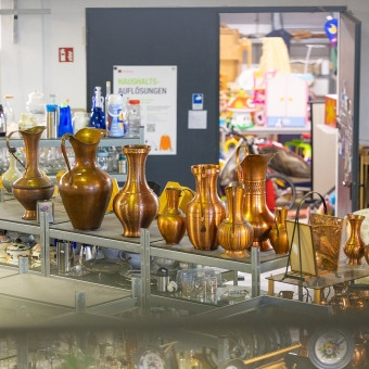 Verschiedene antike, glänzende Kupferkannen und Vasen in verschiedenen Größen und Formen, die auf Metallregalen in einem Secondhand-Laden ausgestellt sind. Im Hintergrund ist ein Schild mit der Aufschrift „Haushaltsauflösungen“ sowie ein Durchgang zu einem weiteren Raum zu sehen.