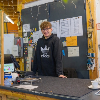 Ein junger Mann mit Brille und lockigem Haar steht lächelnd hinter einem Tresen. Er trägt einen schwarzen Kapuzenpullover mit einem großen weißen Adidas-Logo auf der Vorderseite. Im Hintergrund ist eine dunkle Wand mit verschiedenen Aushängen, Zetteln und Lichterketten zu sehen. Auf dem Tresen befinden sich eine Kasse sowie eine weiße Kaffeetasse.